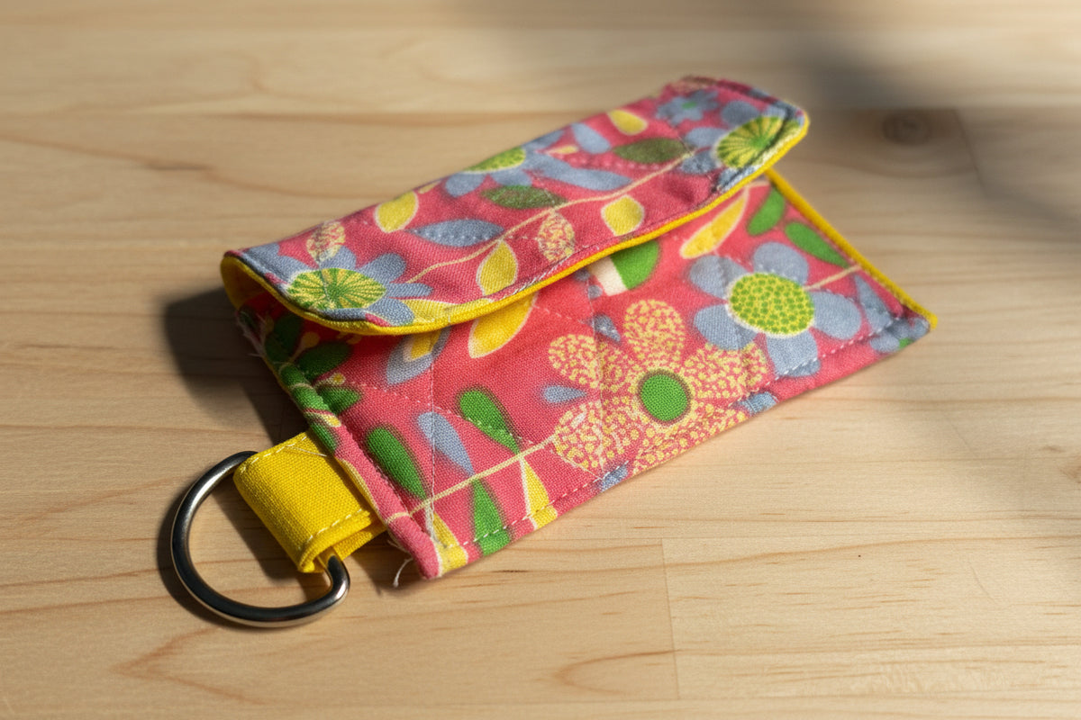 A small fabric pouch with a colorful floral pattern and a yellow loop, resting on a pink cutting mat on a wooden table.
